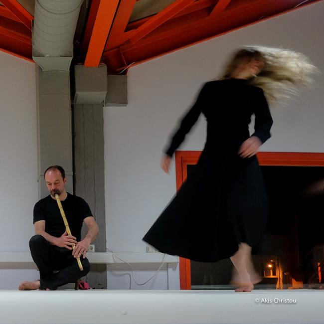 A female dancer is seen vaguely and a musician behind sitting playing the ney in a white attic with red ceiling