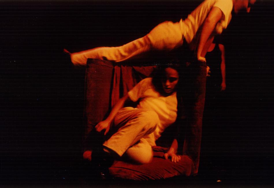 two performers on a red chair, the one sitting and the other on top of it with the body extended diagonally